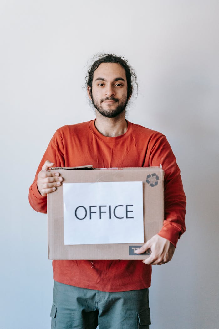 Smiling young man holding a cardboard box labeled 'Office', implying relocation.
